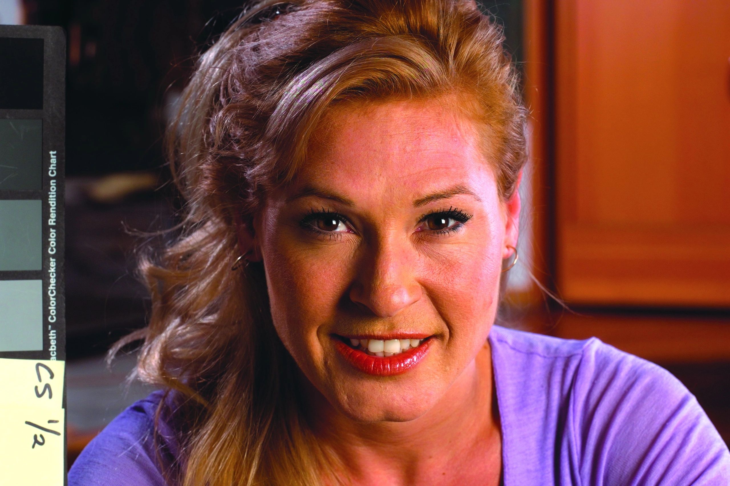 Close-up portrait of a smiling woman with blonde hair wearing a purple shirt, indoors with soft lighting.
