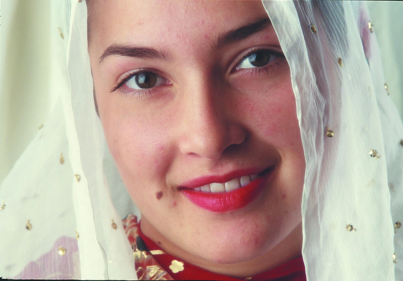 Close-up of a smiling woman with a veil adorned with small golden beads, wearing red lipstick and a red and gold outfit.