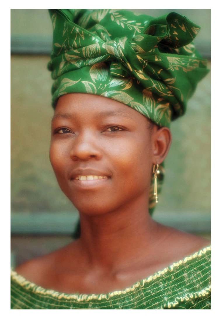 A woman wearing a green patterned headwrap and matching top, with subtle makeup and gold earrings, looks slightly off-camera with a soft expression.