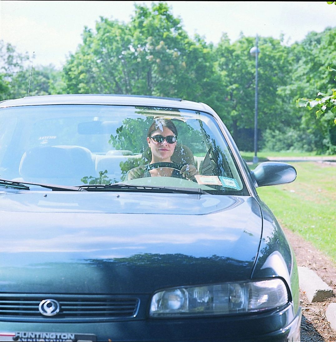 Woman wearing sunglasses, sitting in the driver's seat of a dark sedan, parked in a leafy outdoor area.