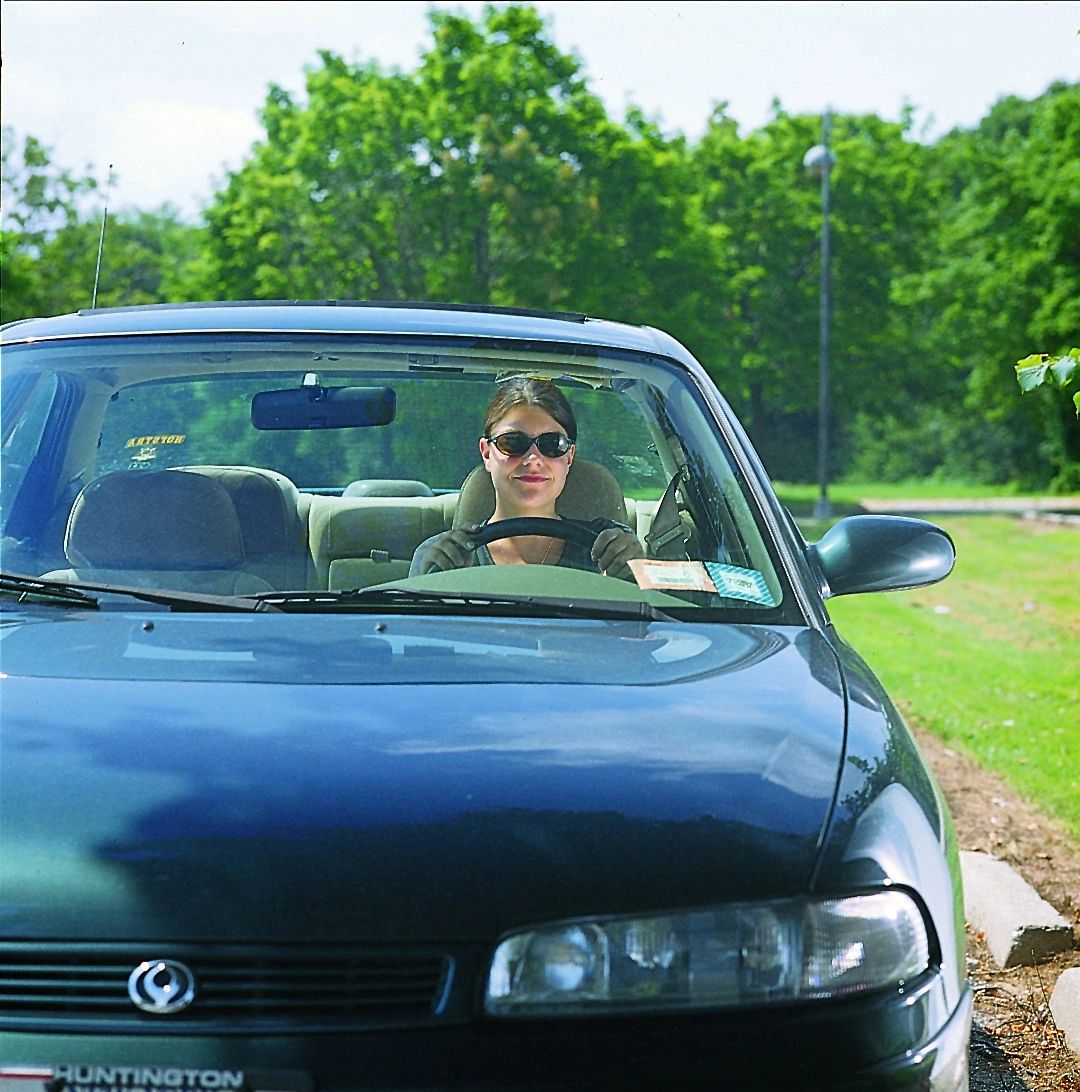 A woman wearing sunglasses sits in the driver's seat of a blue sedan parked outdoors on a sunny day.