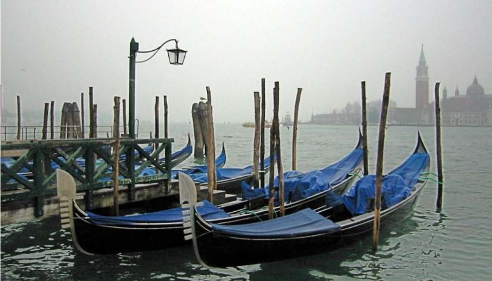 Gondolas covered with blue tarps docked at a misty pier in venice with san giorgio maggiore church in the background.